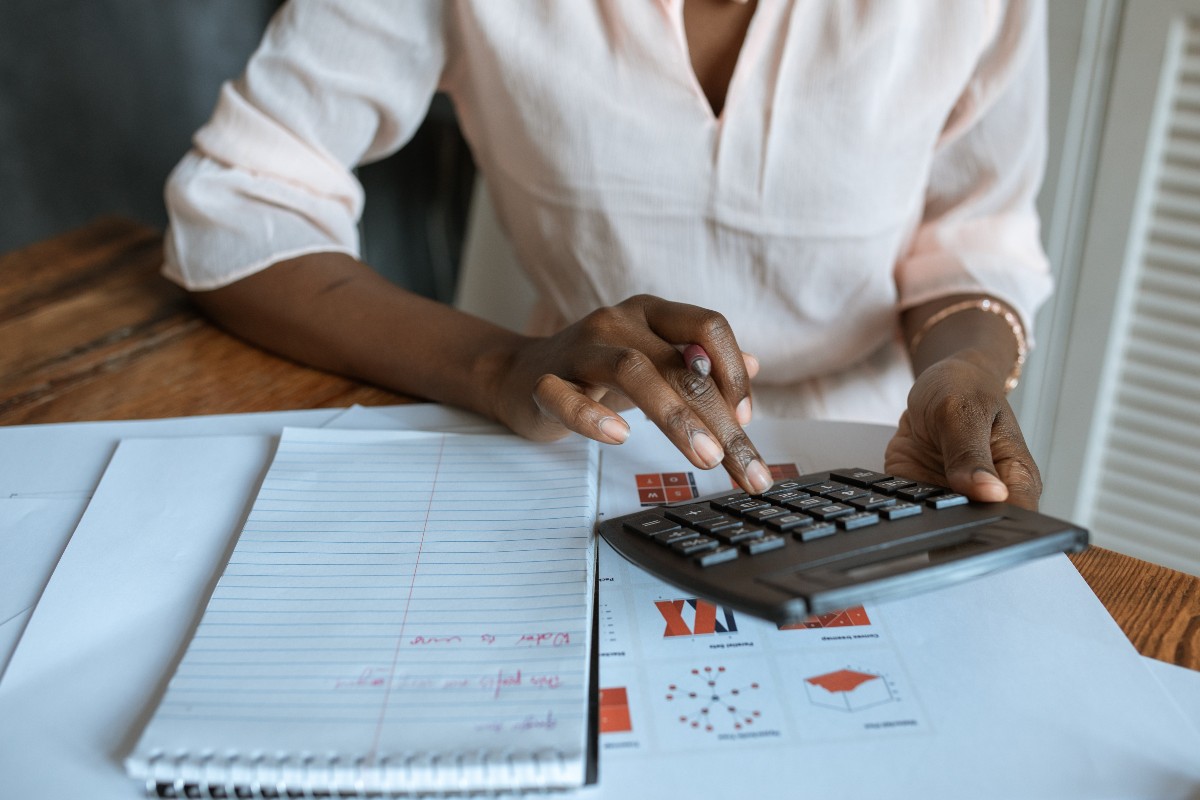 engenharia-de-producao-grade-curricular-carreira-e-salario-Mulher negra utiliza uma calculadora, em frente a uma mesa com um bloco de anotações e uma folha com algumas ilustrações técnicas.