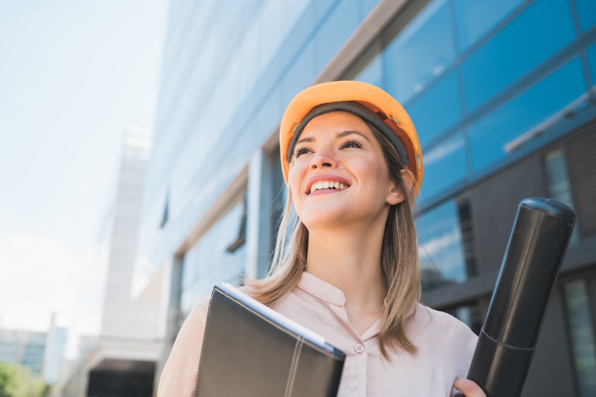 quanto-ganha-um-engenheiro-de-producao-Mulher loira de capacete laranja, com uma pasta e uma bolsa de projeto nas mãos, olha para o horizonte e sorri em frente a um prédio envidraçado.